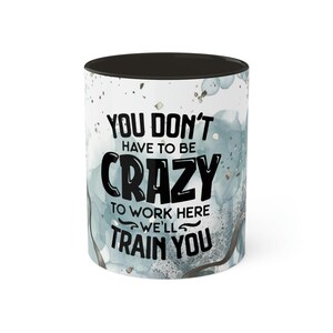 May include: White ceramic coffee mug with a black interior and rim. The mug features the text "YOU DON'T HAVE TO BE CRAZY TO WORK HERE WE'LL TRAIN YOU" in black. The mug has a watercolor-style design in shades of blue and gray.