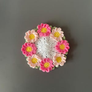 May include: A handmade crochet coaster featuring a central white circle surrounded by a ring of colorful flowers. The flowers have pink and peach petals with yellow centers, and green leaves. This decorative coaster is perfect for adding a touch of handmade charm to any home.