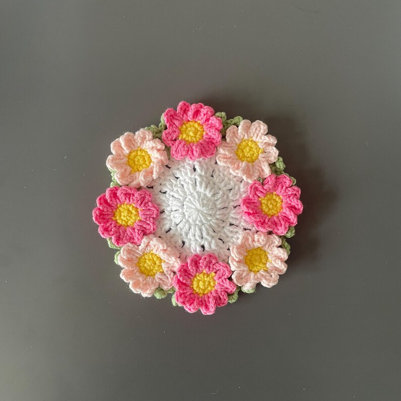 May include: A handmade crochet coaster featuring a central white circle surrounded by a ring of colorful flowers. The flowers have pink and peach petals with yellow centers, and green leaves. This decorative coaster is perfect for adding a touch of handmade charm to any home.