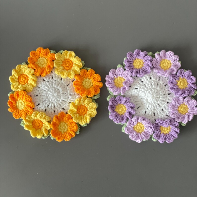 May include: Two handmade crochet coasters. One coaster features a ring of orange and yellow flowers around a white center. The other coaster has a ring of purple flowers with yellow centers around a white center. Both are on a gray surface.