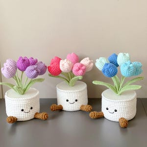 May include: Three crochet flower arrangements in white pots with smiling faces. The flowers are purple, pink, and blue, with green stems and leaves. Each pot has two brown, bean-shaped accents at the base. The background is a neutral color.