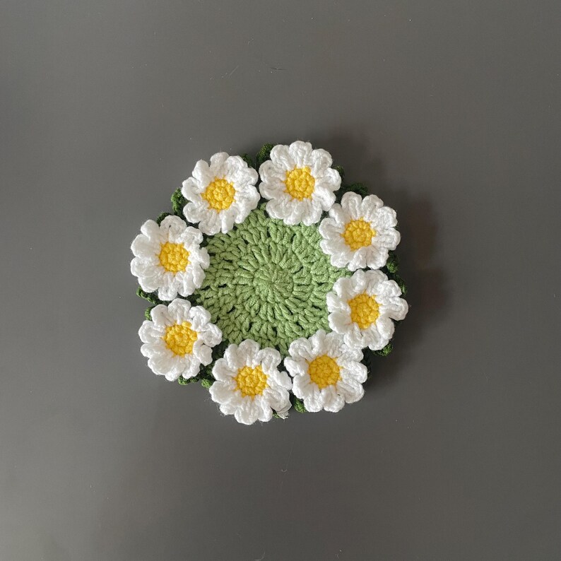 May include: A handmade crochet coaster featuring a daisy design. The coaster has a green center surrounded by white daisy petals with yellow centers. The coaster is round and sits on a gray surface.