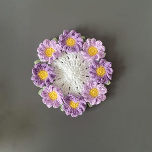 May include: A handmade crochet coaster featuring a white center surrounded by purple flowers with yellow centers and green leaves. The coaster is circular and rests on a gray surface. This item is perfect for home decor.