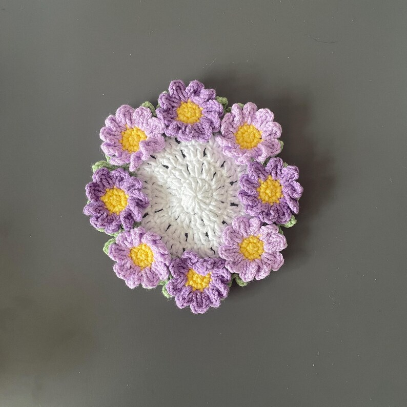 May include: A handmade crochet coaster featuring a white center surrounded by purple flowers with yellow centers and green leaves. The coaster is circular and rests on a gray surface. This item is perfect for home decor.