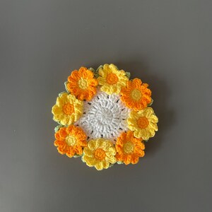 May include: A handmade crochet coaster featuring a central white circle surrounded by a ring of yellow and orange flowers. The coaster is made of yarn and has a floral design, perfect for home decor.