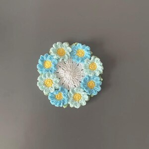 May include: A handmade crochet coaster featuring a daisy design. The coaster has a white center surrounded by light blue and yellow daisies. The coaster is approximately 4 inches in diameter.