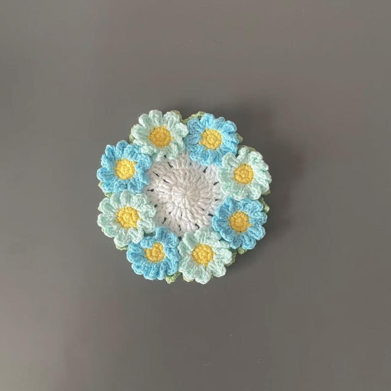 May include: A handmade crochet coaster featuring a daisy design. The coaster has a white center surrounded by light blue and yellow daisies. The coaster is approximately 4 inches in diameter.