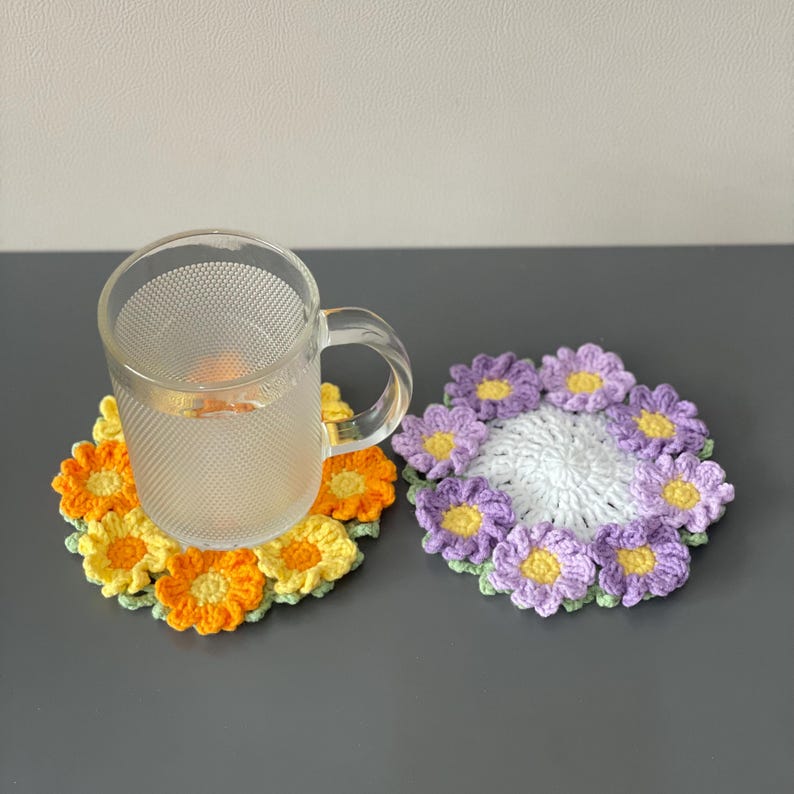 May include: Two handmade crochet coasters in the shape of flowers. One coaster features orange and yellow petals, while the other has purple petals. A clear glass mug with water sits on the orange and yellow coaster.