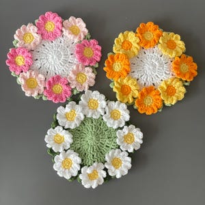May include: Three handmade crochet coasters in a floral design. One coaster features pink and yellow flowers, another has orange and yellow flowers, and the third has white and yellow flowers. Each coaster has a white or green center.