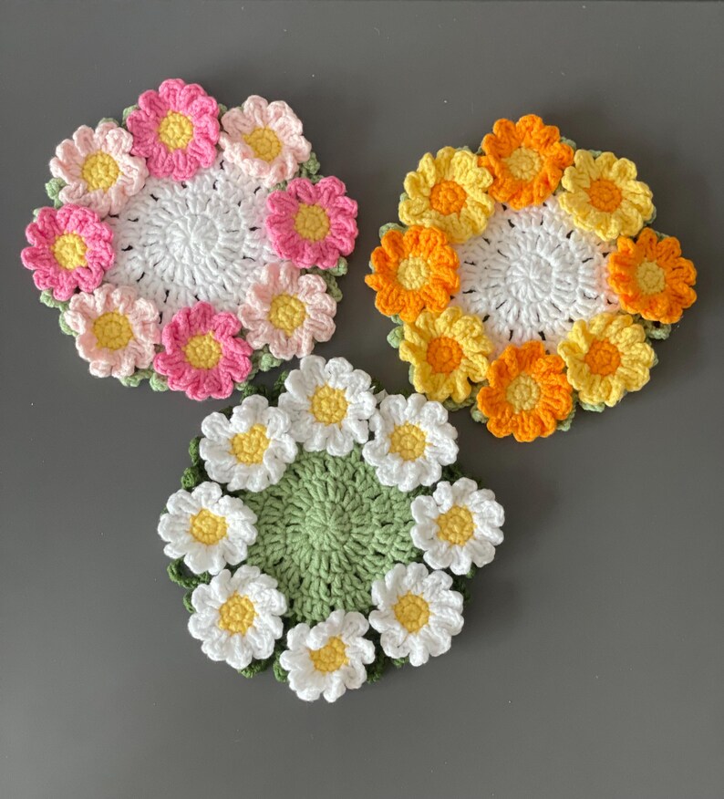 May include: Three handmade crochet coasters in a floral design. One coaster features pink and yellow flowers, another has orange and yellow flowers, and the third has white and yellow flowers. Each coaster has a white or green center.