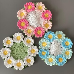 May include: Three handmade crochet coasters in a daisy flower design. One coaster features white and yellow daisies on a green base. Another has pink and yellow daisies. The third has blue and yellow daisies.