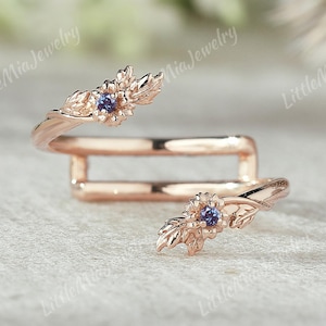 Puede incluir: Un anillo de oro rosa con detalles florales y de hojas. El anillo presenta dos diseños de flores con una pequeña gema azul redonda en el centro. El diseño del anillo envuelve el dedo. El texto "Little Mia Jewelry" está en el fondo.