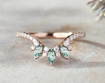Può includere: Una delicata fede nuziale in oro rosa con un design curvo. La fascia è adornata con piccoli diamanti e tre pietre preziose verdi più grandi a forma di marquise.