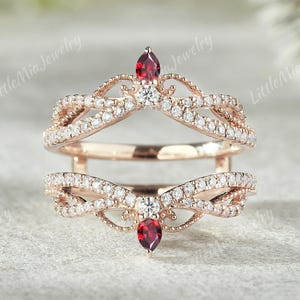 May include: A rose gold ring guard featuring two bands connected by a bridge. The design includes small white pavé stones, two central round white stones, and two pear-shaped red gemstones at the top and bottom points of the ring structure.