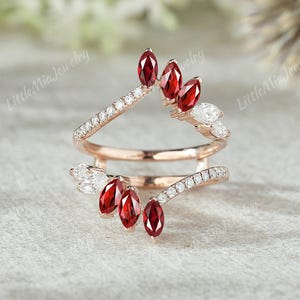 Puede incluir: Un anillo de oro rosa con un diseño único. El anillo presenta una serie de piedras preciosas rojas con corte marquesa y pequeñas piedras preciosas claras y redondas. El anillo tiene un diseño de caña dividida, con las piedras preciosas dispuestas en un patrón floral.