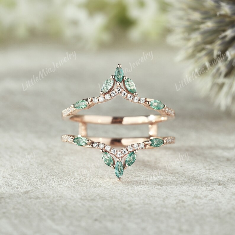 May include: A delicate rose gold ring enhancer with a V-shaped design. The enhancer features a row of small round diamonds and a row of marquise-shaped green gemstones.