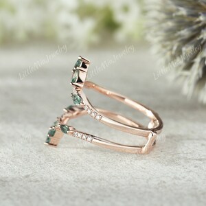 May include: A rose gold ring with a double band design. The top band features a row of small diamonds and five green gemstones. The bottom band is plain.