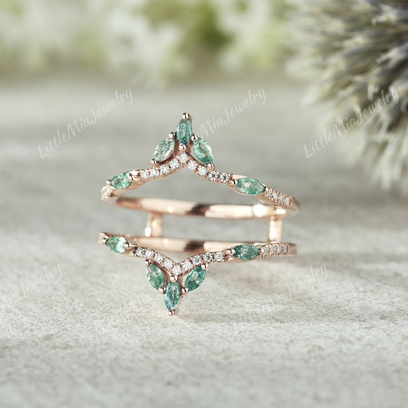 May include: A rose gold ring with a delicate design featuring a row of small diamonds and marquise-shaped green gemstones. The ring is designed to be worn as a wedding band enhancer.