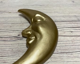 Op de afbeelding: Een goudkleurige halve maan sculptuur met een lachend gezicht. De maan heeft een gladde, metallic afwerking en staat op een lichtgekleurd houten oppervlak. De sculptuur is een decoratief item.