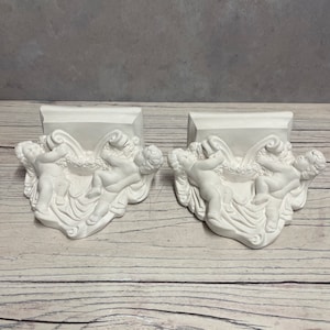 May include: A pair of white plaster wall brackets featuring cherubs holding a floral wreath. The brackets are designed to be mounted on a wall and can be used to display decorative items.