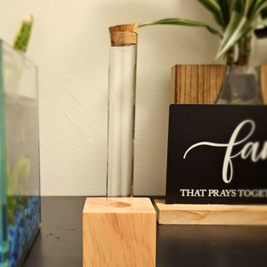 May include: A small, clear glass test tube with a cork stopper sits on a wooden block. The block is a light brown color and has a smooth surface. The test tube is empty and appears to be made of glass. The test tube is sitting on a black surface. The background of the image is a light brown wall.