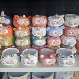 May include: A collection of ceramic planters in various colors, shaped like pigs and other animals. The planters have cartoonish faces and are arranged on a shelf. Some have unicorn horns or bear ears.