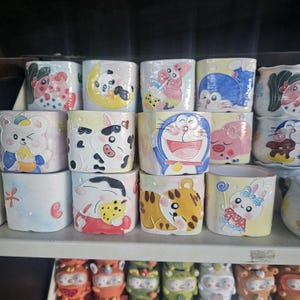 May include: A collection of 15 colorful ceramic planters featuring various cartoon characters, including a panda, a tiger, a cat, and a cartoon robot. The planters are all square shaped and have a glossy finish.