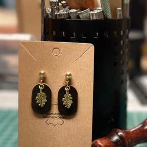 May include: A pair of brown leather earrings with gold leaf accents. The earrings are displayed on a brown card with the text "Flatlands Leathercraft".