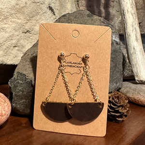May include: A pair of black leather earrings with gold chains and gold ball accents. The earrings are shaped like half circles and are displayed on a brown card with the text "Flatlander Leathercraft".