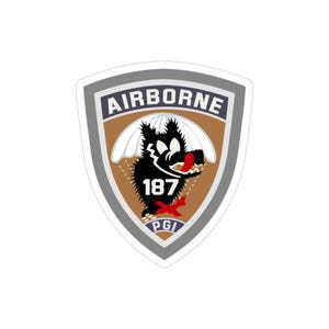Può includere: Un adesivo con un design a scudo con la scritta "AIRBORNE" in alto. Lo scudo ha uno sfondo marrone con una grafica di lupo nero, il numero "187" e le lettere "PGI". Il lupo è sotto un paracadute bianco.