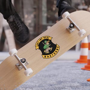 Può includere: Uno skateboard con un adesivo raffigurante un soldato dei cartoni animati in verde e nero, con la scritta "327 INF REG BASTOGNE" in giallo. Lo skateboard ha una finitura in legno chiaro e hardware argentato.