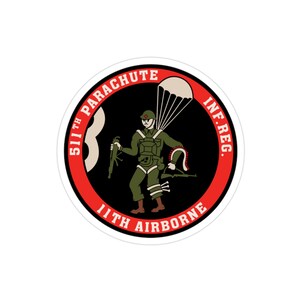 Op de afbeelding: Een ronde sticker met een militair embleem. Het ontwerp toont een soldaat met een parachute en geweer, en de tekst "511th Parachute Inf. Reg. 11th Airborne" in rood en wit.