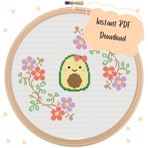 May include: Cross stitch pattern featuring a cute avocado with a pink bow and green leaves, surrounded by pink and purple flowers.  The pattern is displayed in a wooden embroidery hoop.