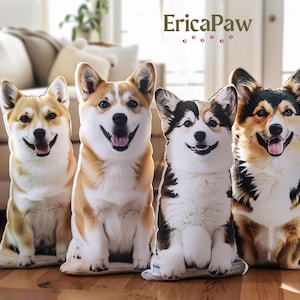 May include: Four plush dog pillows featuring different corgi breeds. The pillows are white with brown, black, and tan markings. The pillows are arranged in a row on a wooden floor. The text "EricaPaw" is visible in the background.