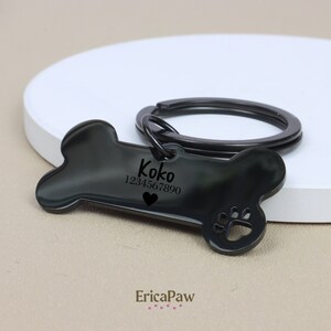 Op de afbeelding: Zwarte, botvormige hondenhanger met de naam "Koko" en het telefoonnummer "1234567890" erop gegraveerd. De hanger heeft een klein hartje en een pootprint.