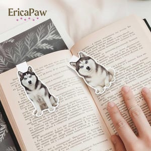 Può includere: Due segnalibri a forma di cane husky sono posizionati sulle pagine di un libro aperto. I segnalibri sono bianchi con un'immagine realistica di un cane husky. Il libro ha testo in lingua straniera. Il marchio "EricaPaw" è in alto.