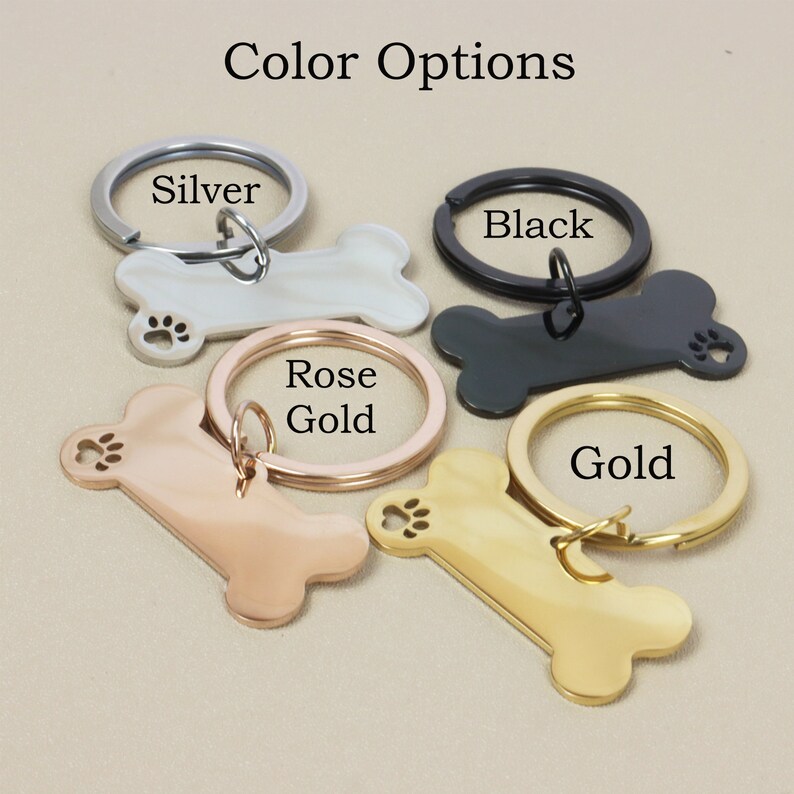 Op de afbeelding: Vier sleutelhangers in de vorm van een hondenbot in verschillende kleuren: zilver, zwart, ros&eacute; goud en goud. Elke sleutelhanger heeft een klein gegraveerd pootje.