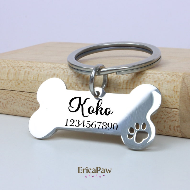 Op de afbeelding: Zilveren hondenpenning in de vorm van een bot met de naam "Koko" in zwarte gravure en het nummer "1234567890". De penning heeft een uitsparing in de vorm van een poot en een zilveren ring.
