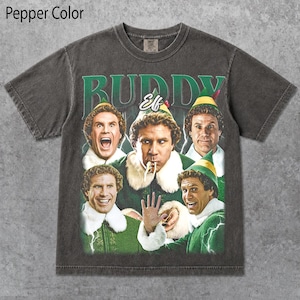 Puede incluir: Una camiseta de manga corta color pimienta con un estampado gráfico del personaje Buddy the Elf en varias poses. El texto BUDDY está impreso en letras verdes grandes en la parte superior, con la palabra Elf en letra blanca debajo.