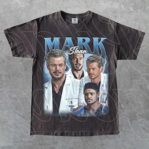 May include: Dark brown t-shirt with a vintage-style graphic of Mark Sloan from Grey's Anatomy. The design includes multiple images of the character with the name "MARK Sloan" in blue text. The shirt has a worn, distressed appearance.