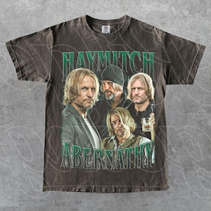 Puede incluir: Camiseta marrón con un gráfico de un hombre en cuatro poses diferentes. El texto "HAYMITCH ABERNATHY" está impreso en verde arriba y abajo de las imágenes. La camiseta tiene un aspecto vintage y desgastado.