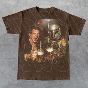 May include: A brown short-sleeve t-shirt featuring a graphic print of a laughing man and a Boba Fett character both holding glass beer mugs. The design shows the man with an open mouth and the character wearing a green and yellow helmet.