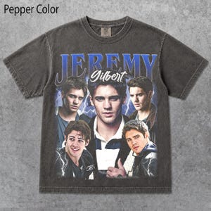 May include: A pepper-colored short-sleeve t-shirt featuring a graphic print of the character Jeremy Gilbert. The design includes the name Jeremy in large blue letters, the name Gilbert in white script, and four photographic portraits of the character.