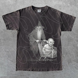 May include: Dark grey t-shirt with a black and white graphic of Gandalf pushing a shopping trolley with Gollum inside. The design is a parody of the Lord of the Rings. The shirt has a vintage, distressed appearance.