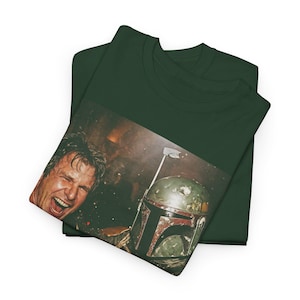 May include: A dark forest green t-shirt featuring a rectangular graphic print. The image displays a close-up of a person with an open-mouthed expression next to a weathered, green and red helmet with a metallic antenna, set against a dark background.