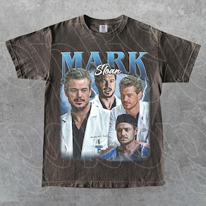 May include: Brown t-shirt with a vintage graphic of Mark Sloan from Grey's Anatomy. The design shows multiple images of the character with the name "MARK Sloan" in a blue, stylised font. The shirt has a worn, distressed appearance.