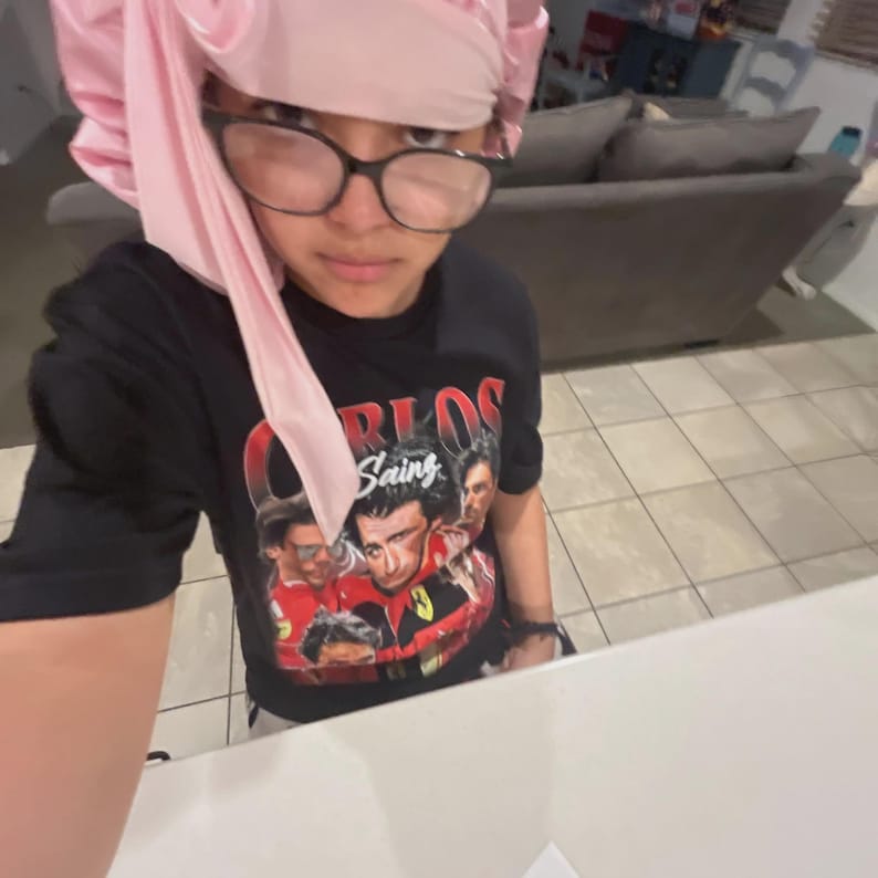 Op de afbeelding: Zwart T-shirt met een afbeelding van Carlos Sainz en andere racefiguren. De persoon draagt een roze satijnen muts en een bril met zwart montuur. De afbeelding is een selfie die binnenshuis is gemaakt.