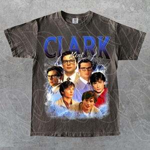 Op de afbeelding: Donkergrijs t-shirt met een afbeelding van meerdere afbeeldingen van Clark Kent, met de naam "CLARK Kent" in blauw. Het ontwerp heeft een vintage, distressed look met bliksemaccenten.