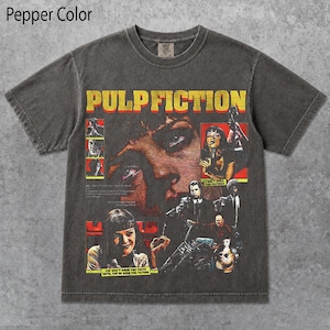 May include: A pepper-colored short-sleeve t-shirt featuring a large graphic print from the movie Pulp Fiction. The design includes the yellow text Pulp Fiction, a large close-up of a face, and several smaller scenes with characters from the film.