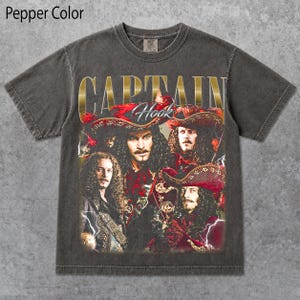 May include: A pepper-colored short-sleeve t-shirt featuring a graphic print of Captain Hook. The design includes the text "CAPTAIN Hook" in gold lettering above four portraits of the character wearing a red pirate hat and ornate clothing.
